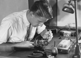 Black-and-white photo of a man sitting on a bench, adjusting a Bolex camera. 